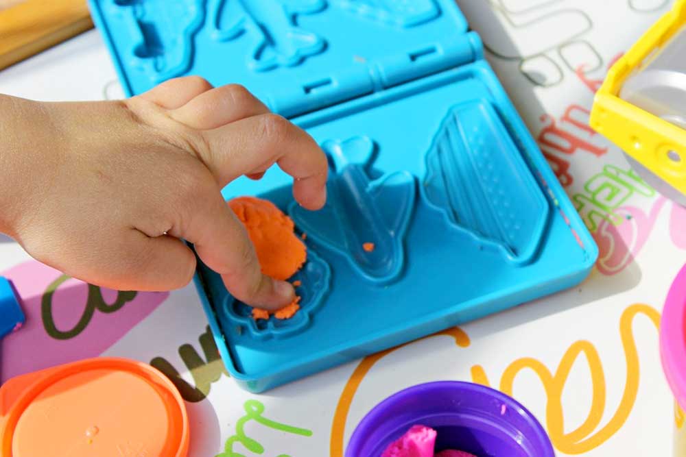 Easter Basket - Child's hand pressing orange play-doh into a blue mold with spaces to make a car, plane, or cruise ship.