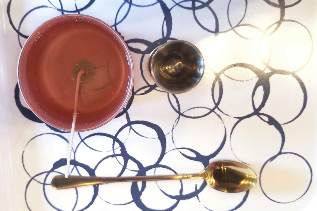 White tray with blue interlocking circles. On the tray is a liquid candle, gold jigger, and gold spoon