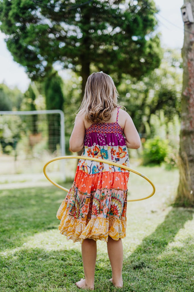 A Girl in Floral Dress Standing on the Green Grass While Doing Hula Hoop