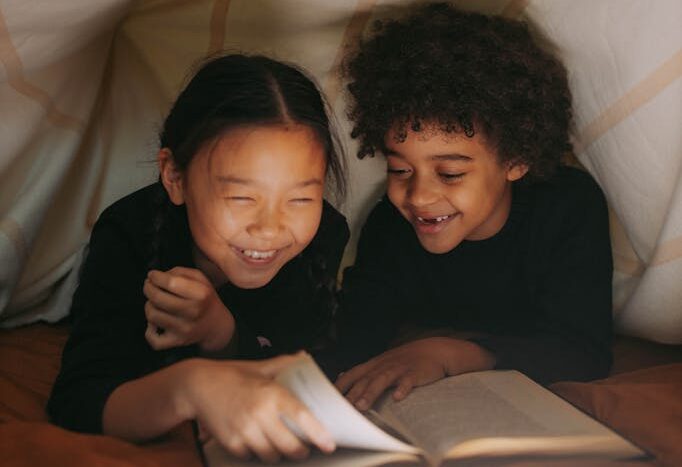 A Young Girl and a Boy Reading a Book while Under the Blanket