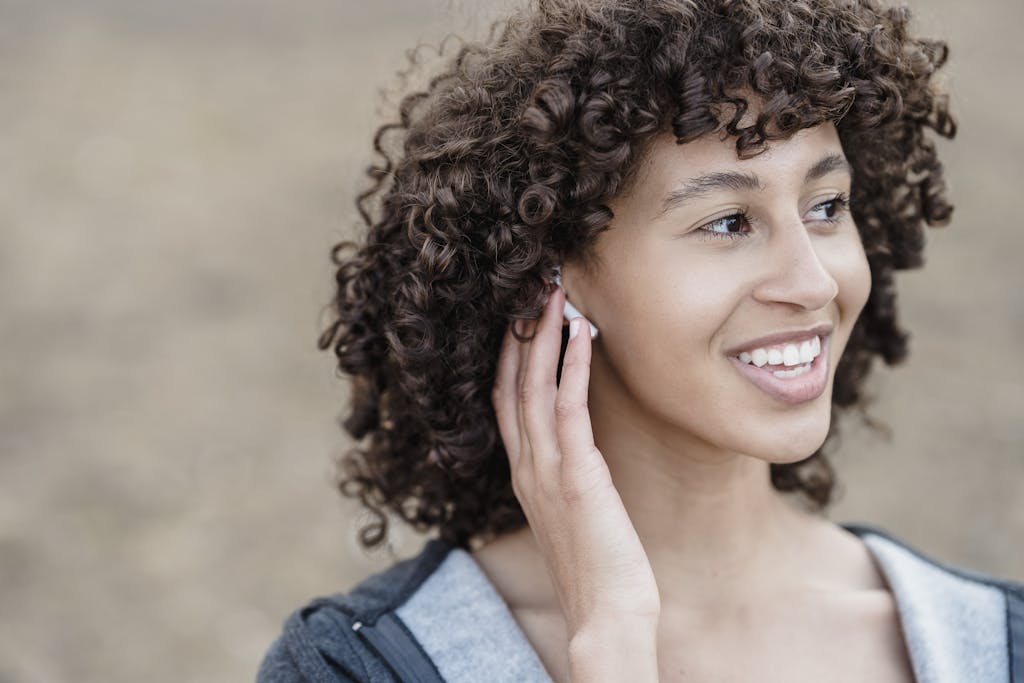 Cheerful ethnic female with black curly hair looking away with smile while listening to songs in wireless earphones on street against blurred background
