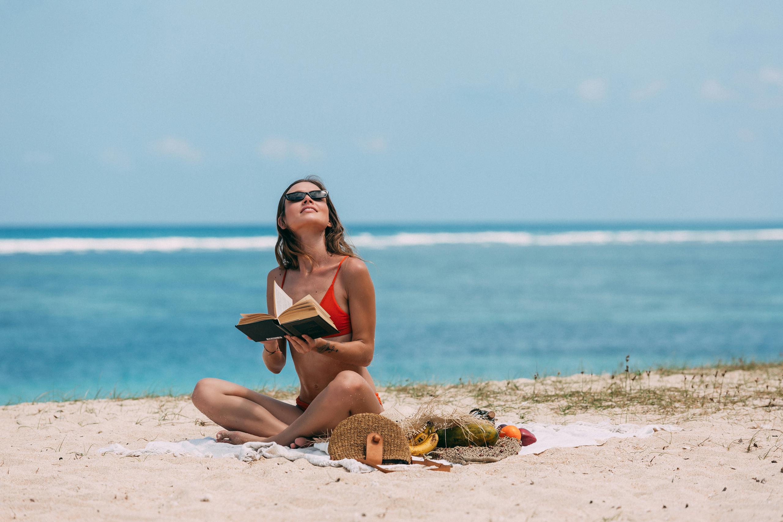 Hot List - Woman in bikini reading a book on a sunny beach, enjoying summer vacation.